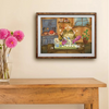 A framed art print depicting a whimsical frog character in a kitchen, styled on a beige wall above a wooden console table with a vase of pink flowers.