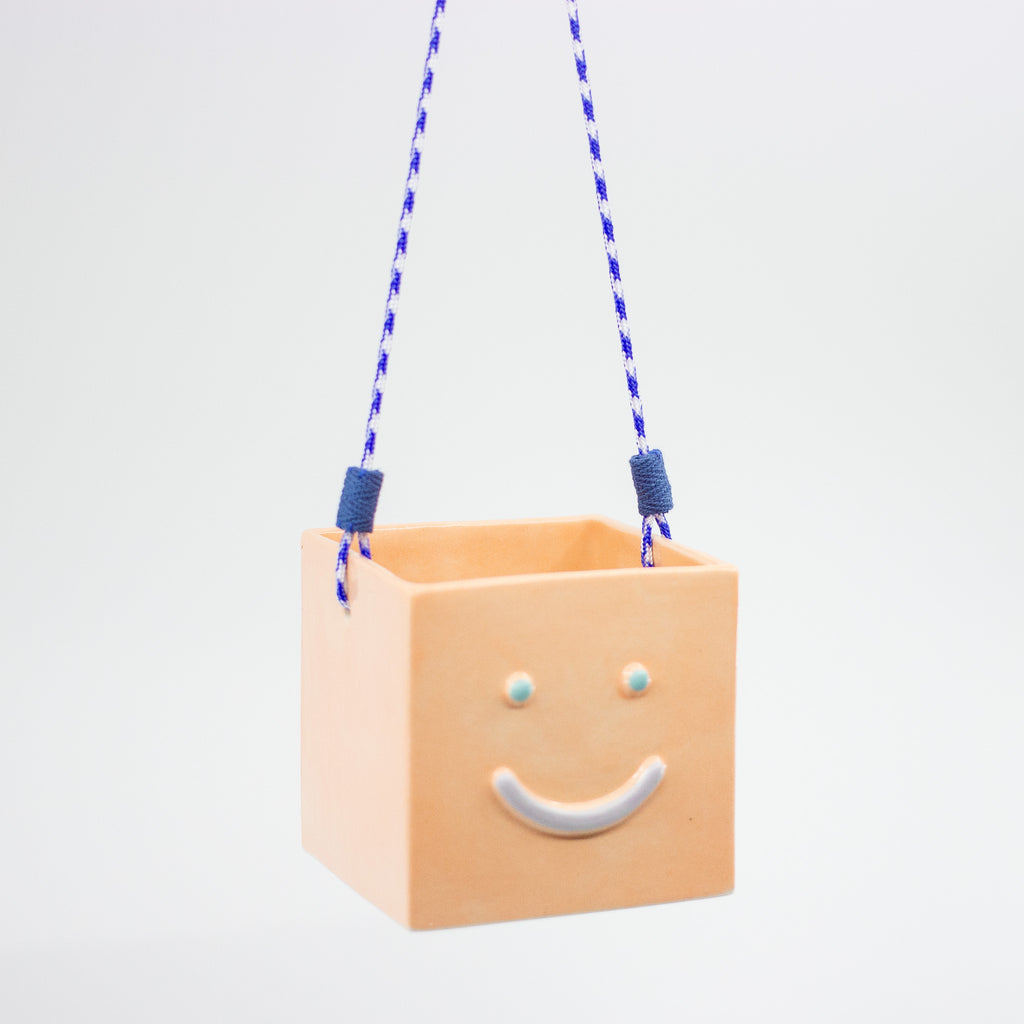 Square ceramic hanging planter with a smiley face and blue swing ropes on a light gray background