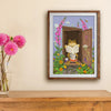 A framed art print depicting a frog reading a book in a garden, hanging on a beige wall above a wooden console table with a vase of pink flowers.