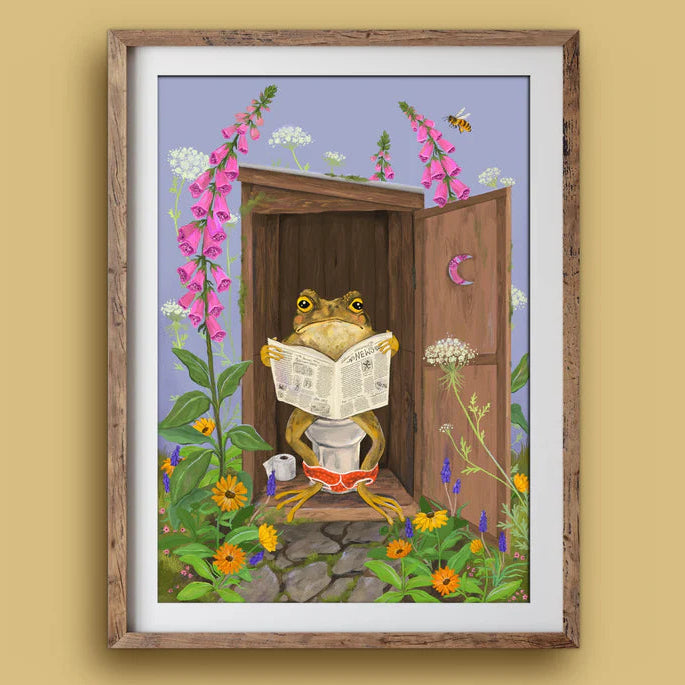 A framed art print featuring a whimsical illustration of a frog sitting on a toilet and reading a newspaper, surrounded by flowers and a wooden door, displayed on a beige wall.