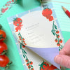 A hand is peeling back a printed paper layer to reveal a layer underneath, showing a tomato-themed shopping list. The top sheet features cute red cartoon tomatoes and tomato vines on a light blue grid background.