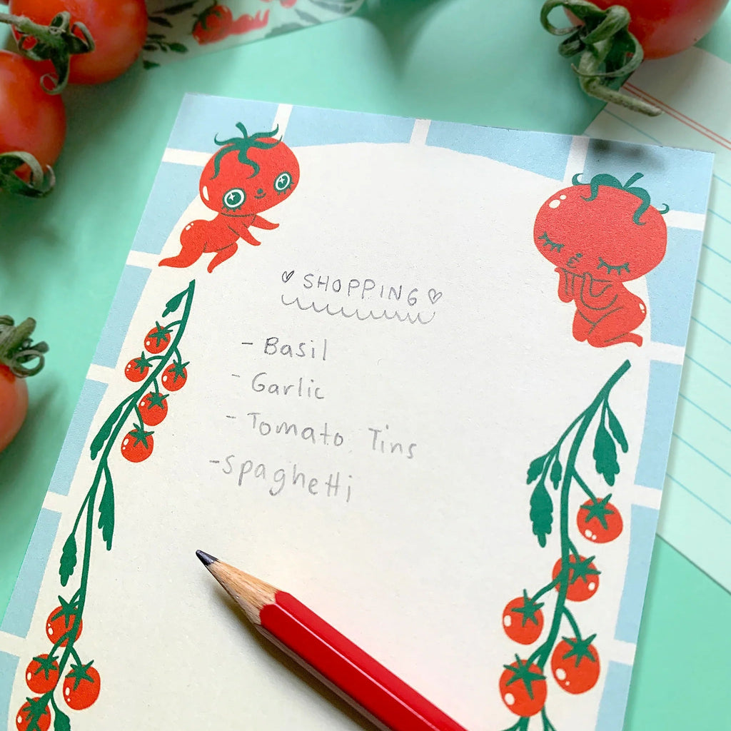 A square notepad with a cute tomato-themed illustration, featuring a shopping list written on it. The notepad is styled on a mint green surface with fresh tomatoes, a red pencil, and patterned tape around it.