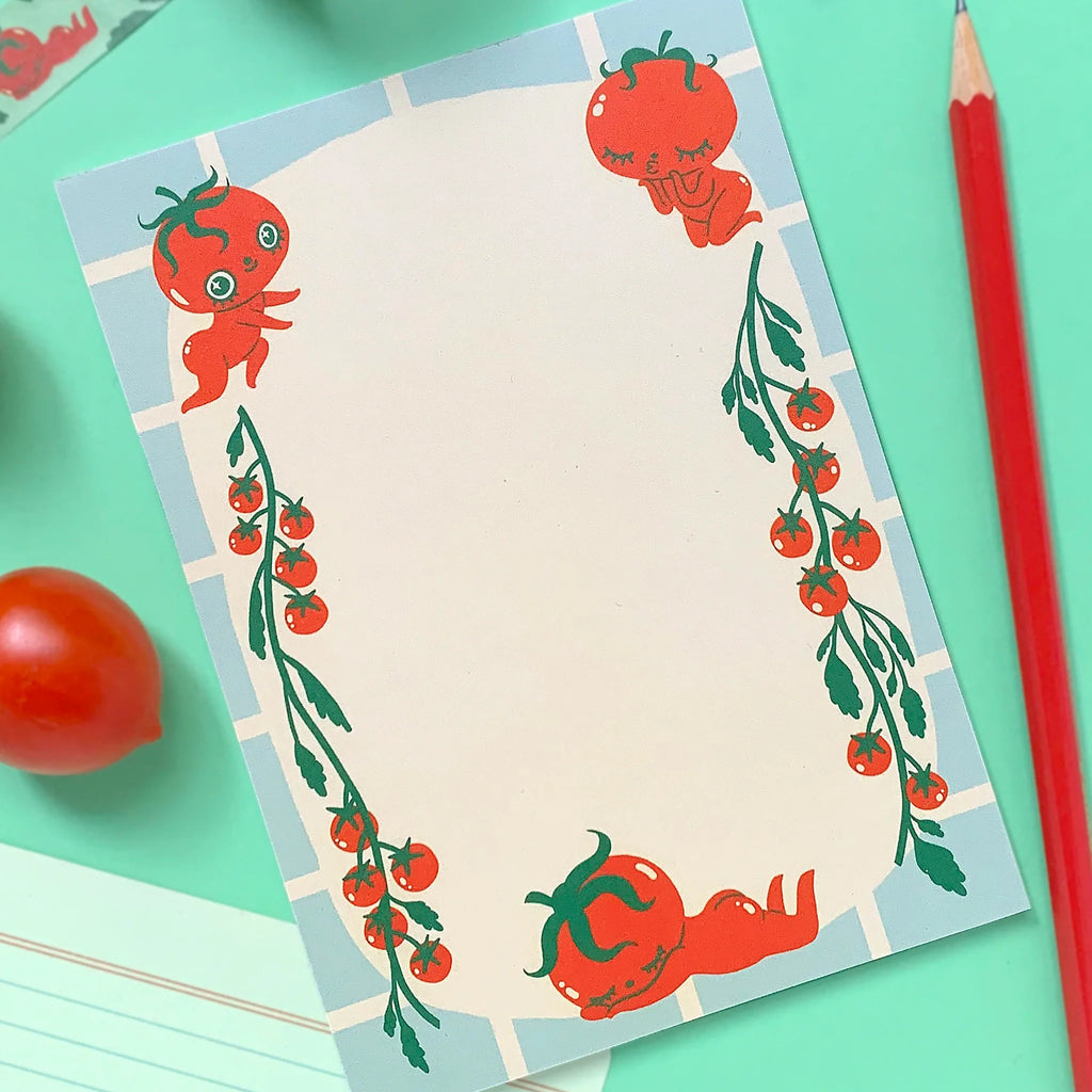 A flat-lay of a notepad with a cute tomato-themed border design, styled on a mint green background with a red pencil, cherry tomatoes, and a roll of patterned tape.