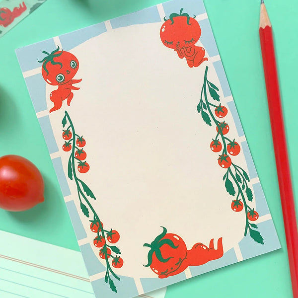 A flat-lay of a notepad with a cute tomato-themed border design, styled on a mint green background with a red pencil, cherry tomatoes, and a roll of patterned tape.