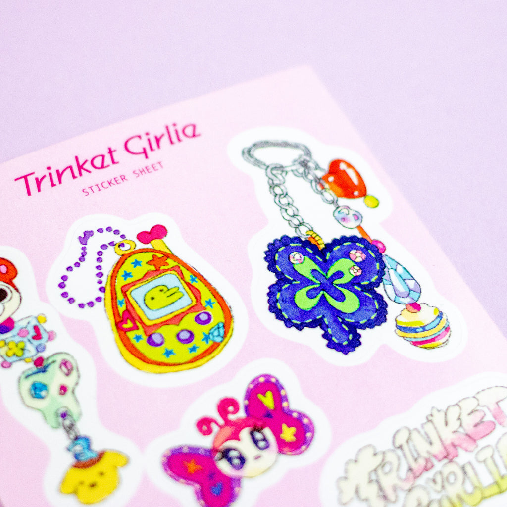 A close-up of a sticker sheet labeled 'Trinket Girlie STICKER SHEET' on a light pink background, showing colourful cartoon-style stickers including a yellow tamagotchi, a blue flower keychain charm, and other playful designs.