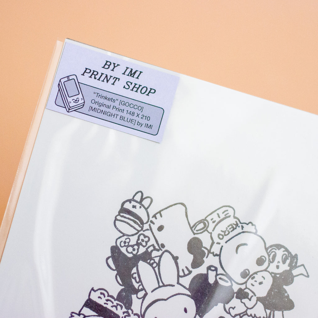 A close-up of a printed sheet corner with a visible label reading 'BY IMI PRINT SHOP' and cartoon characters printed in black on a white surface, set against a peach background.