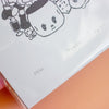 A close-up of an art print featuring a black line-art illustration of a cute character on a white background, with handwritten text including 'TRINKETS', '55/60'. A peach-coloured surface is visible at the bottom right.