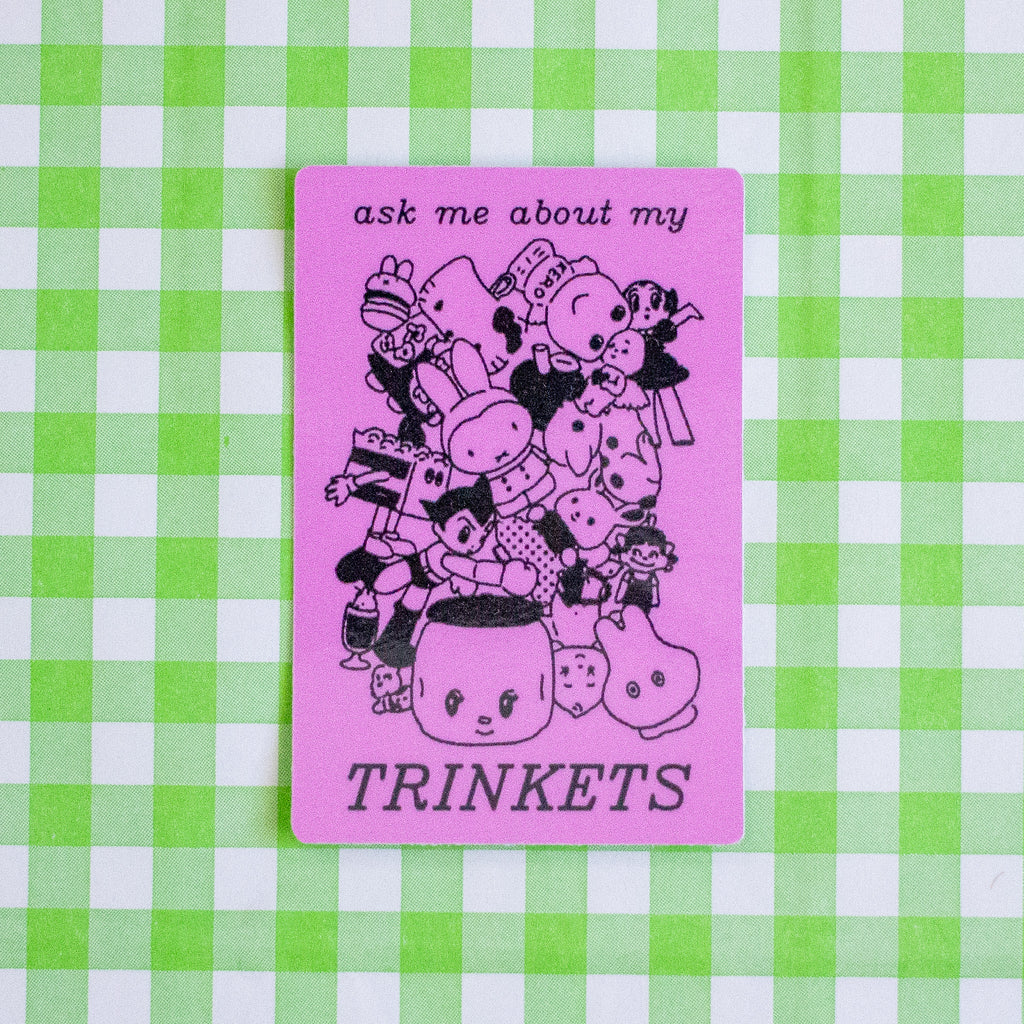 A rectangular pink sticker featuring black line-art illustration of cartoon illustrations and the text 'ask me about my TRINKETS', placed on a green and white checkered background.