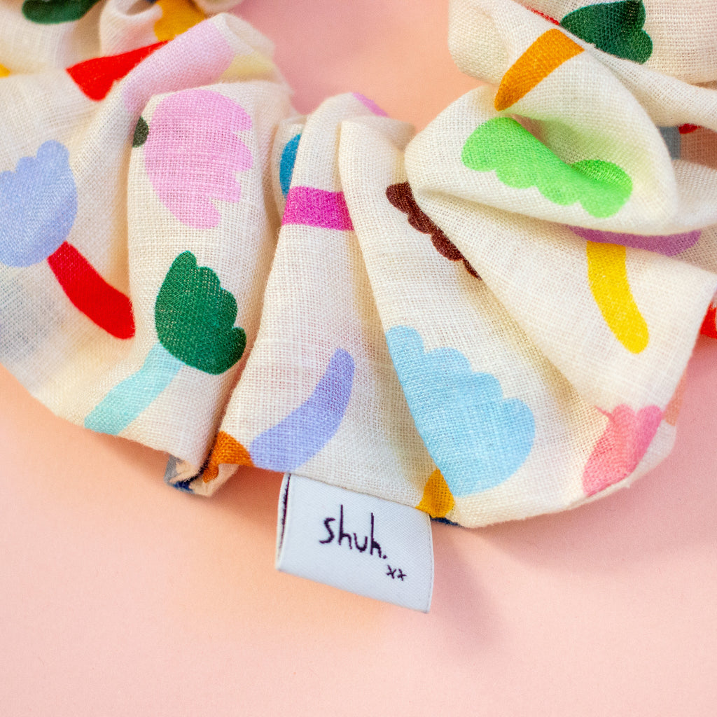 Colourful floral scrunchie on a peach background, featuring a 'shuh' label.