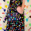 A person wearing a black dress with a colourful abstract tulip-like print carries a matching black crossbody bag with the same print. The background is a matching patterned textile.