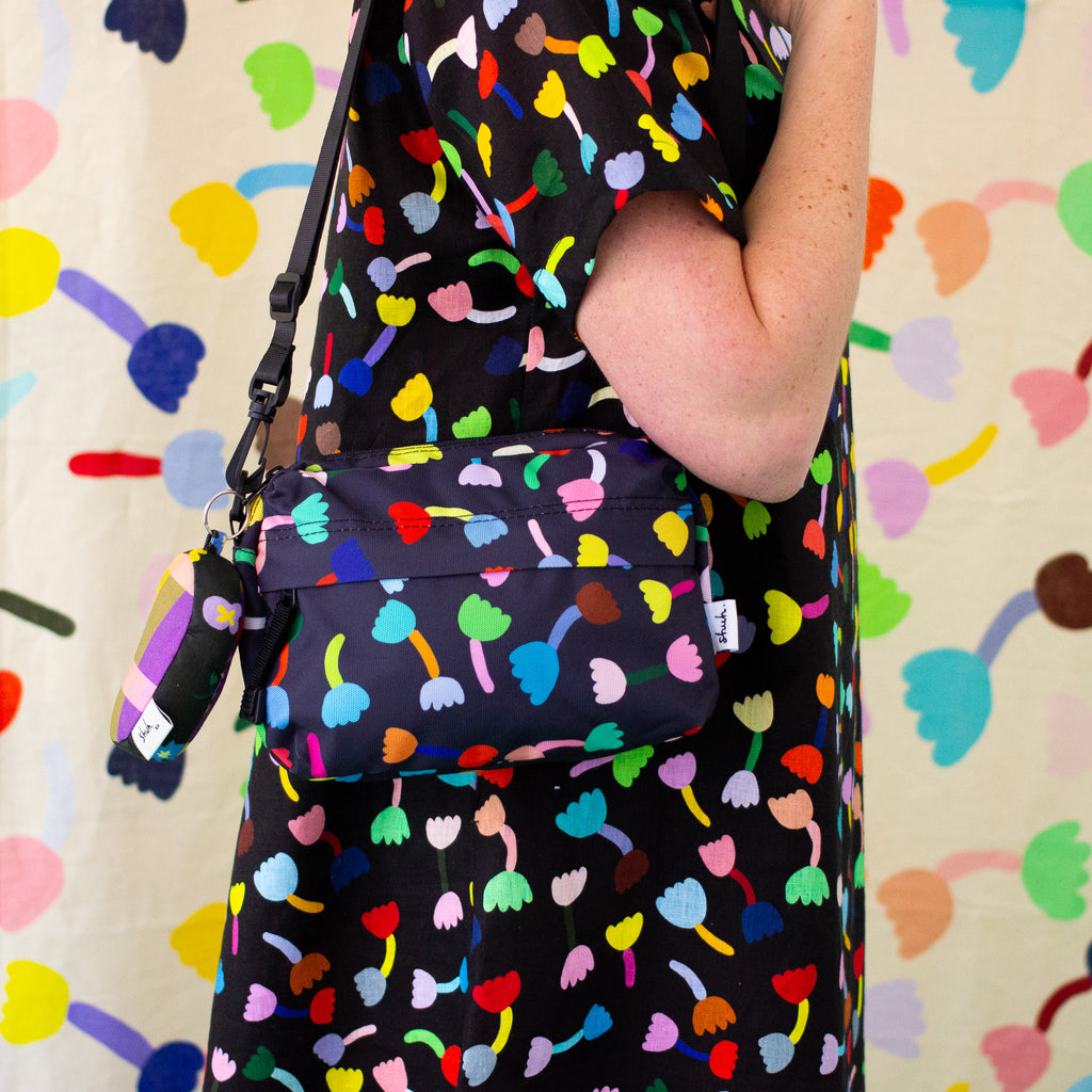 A person wearing a black dress with a colourful abstract tulip-like print carries a matching black crossbody bag with the same print. The background is a matching patterned textile.