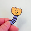A small enamel pin shaped like a smiling flower, held between two fingers against a plain light background.