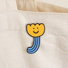 A close-up of a canvas tote bag in a warm off-white colour, featuring a small enamel pin shaped like a smiling yellow flower with a blue stem.