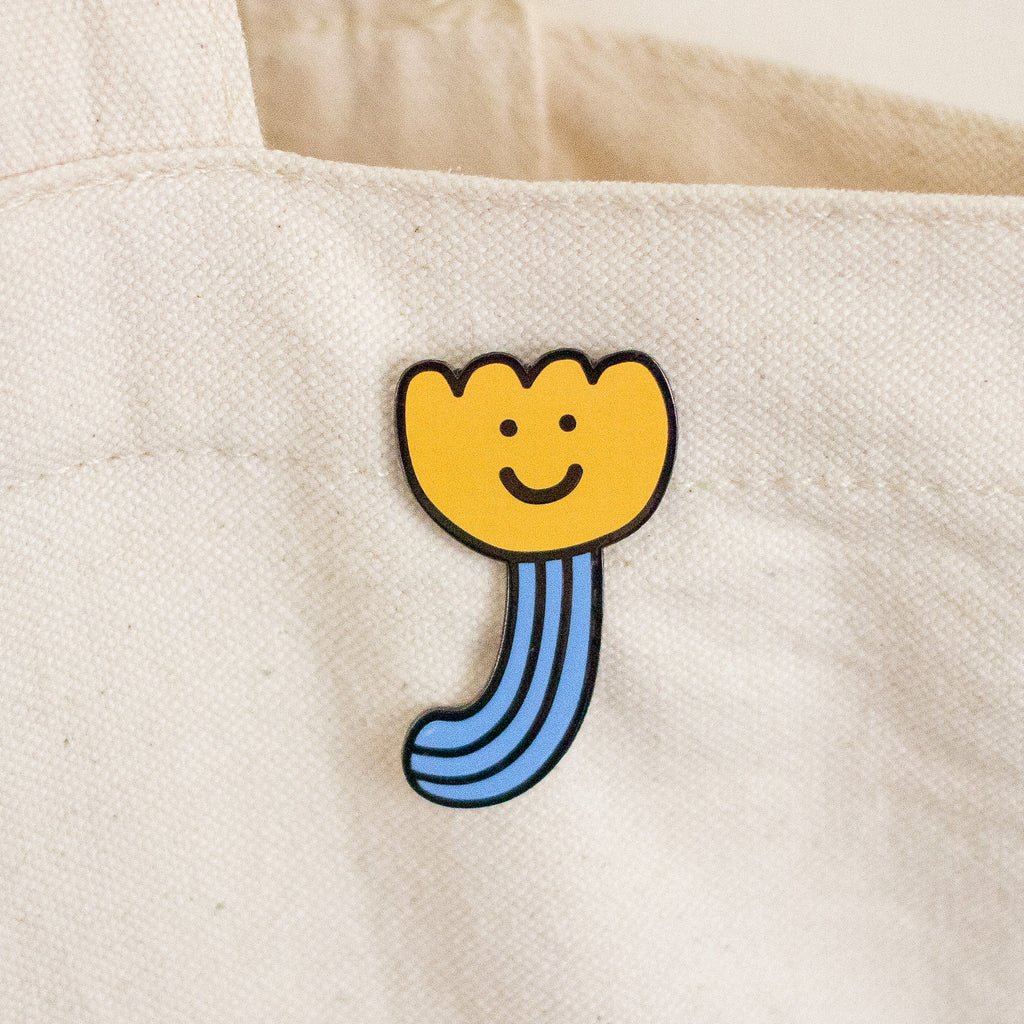 A close-up of a canvas tote bag in a warm off-white colour, featuring a small enamel pin shaped like a smiling yellow flower with a blue stem.