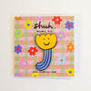 A yellow flower-shaped enamel pin with a smiling face and blue striped stem, mounted on a square pink and beige gingham card with colourful flowers and the text 'shuh Enamel Pin' and 'www.shuhlee.com'.