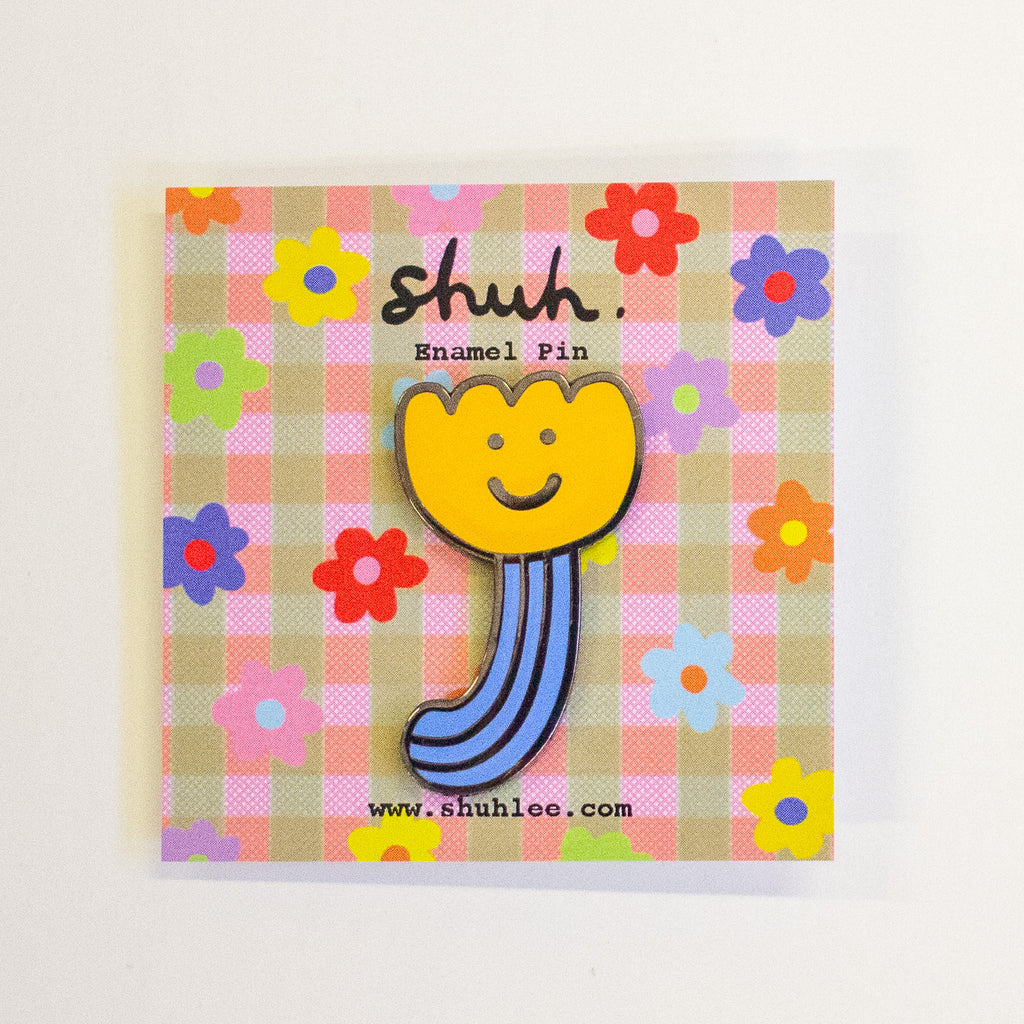 A yellow flower-shaped enamel pin with a smiling face and blue striped stem, mounted on a square pink and beige gingham card with colourful flowers and the text 'shuh Enamel Pin' and 'www.shuhlee.com'.