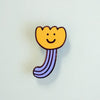 A small enamel pin shaped like a smiling tulip flower, featuring a yellow-orange flower head with a simple black face and a blue striped stem, shown on a plain light background.