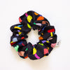 Black fabric scrunchie featuring a playful and colourful floral print with red, yellow, blue, green, and pink elements, displayed in a circular shape on a white background.