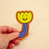 A hand holding a die-cut sticker shaped like a smiling flower with a yellow bloom, orange border, and purple striped stem on a plain beige background.