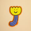 A die-cut sticker featuring a whimsical, cartoon-style flower with a yellow bloom, purple striped stem, and orange border, displayed on a solid beige background.