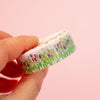 A hand holding a roll of washi tape decorated with a colourful tulip field on a soft pink background.