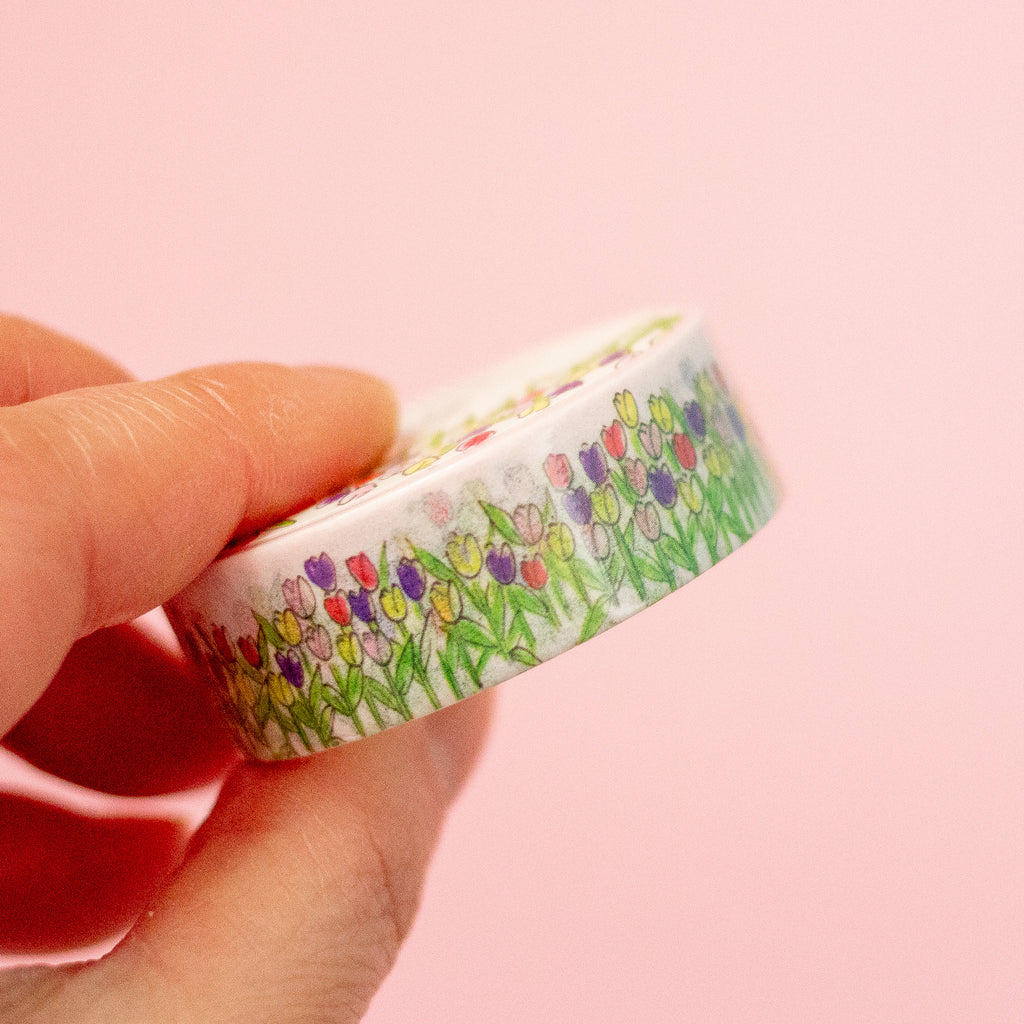 A hand holding a roll of washi tape decorated with a colourful tulip field on a soft pink background.