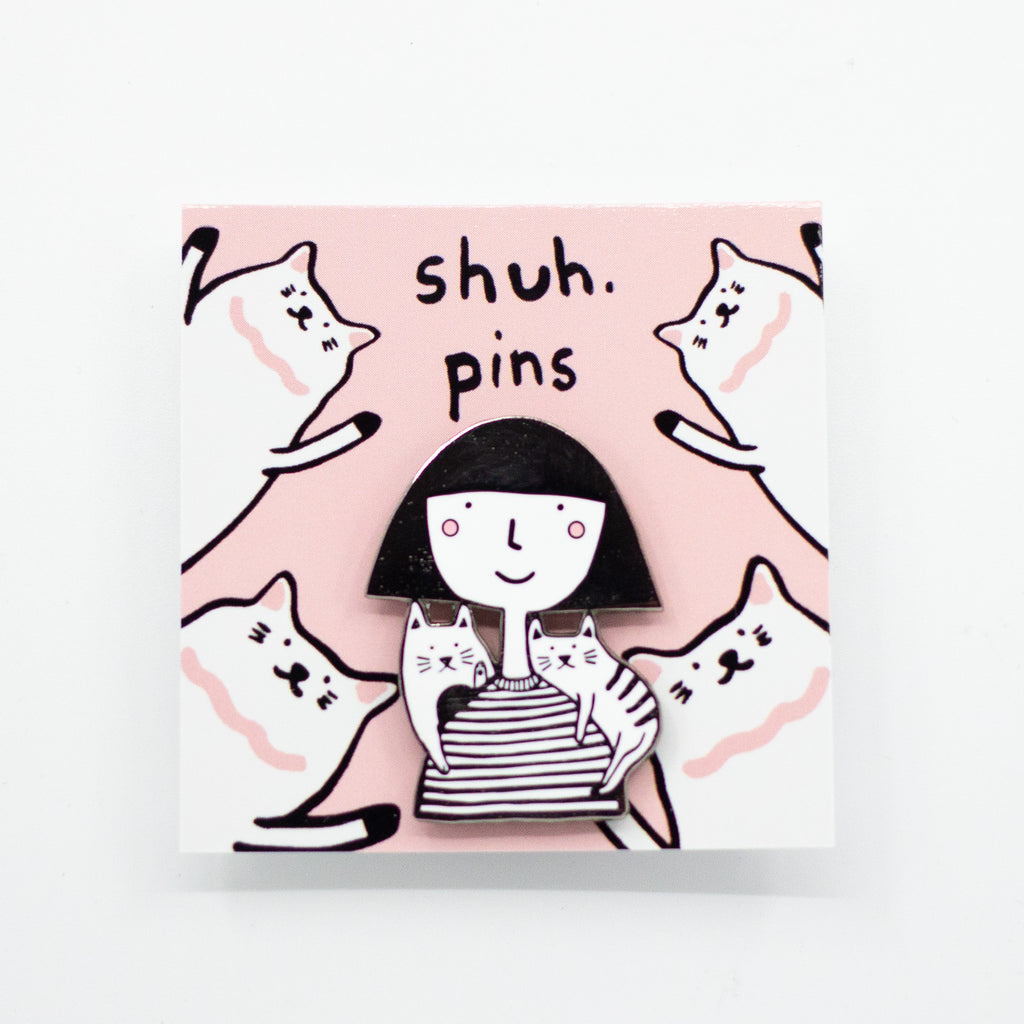 Enamel pin showing two hand-drawn cats sitting on the shoulders of a girl in a striped tee, displayed on a pink backing card with illustrated cats and “shuh. pins” branding.