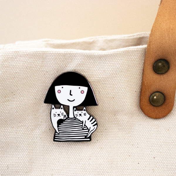 Enamel pin showing two hand-drawn cats sitting on the shoulders of a girl in a striped tee, attached to a calico bag. 