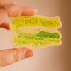 A hand holds a die-cut sticker featuring green ants carrying yellow banner that reads 'STRONGER TOGETHER' on a warm beige background.