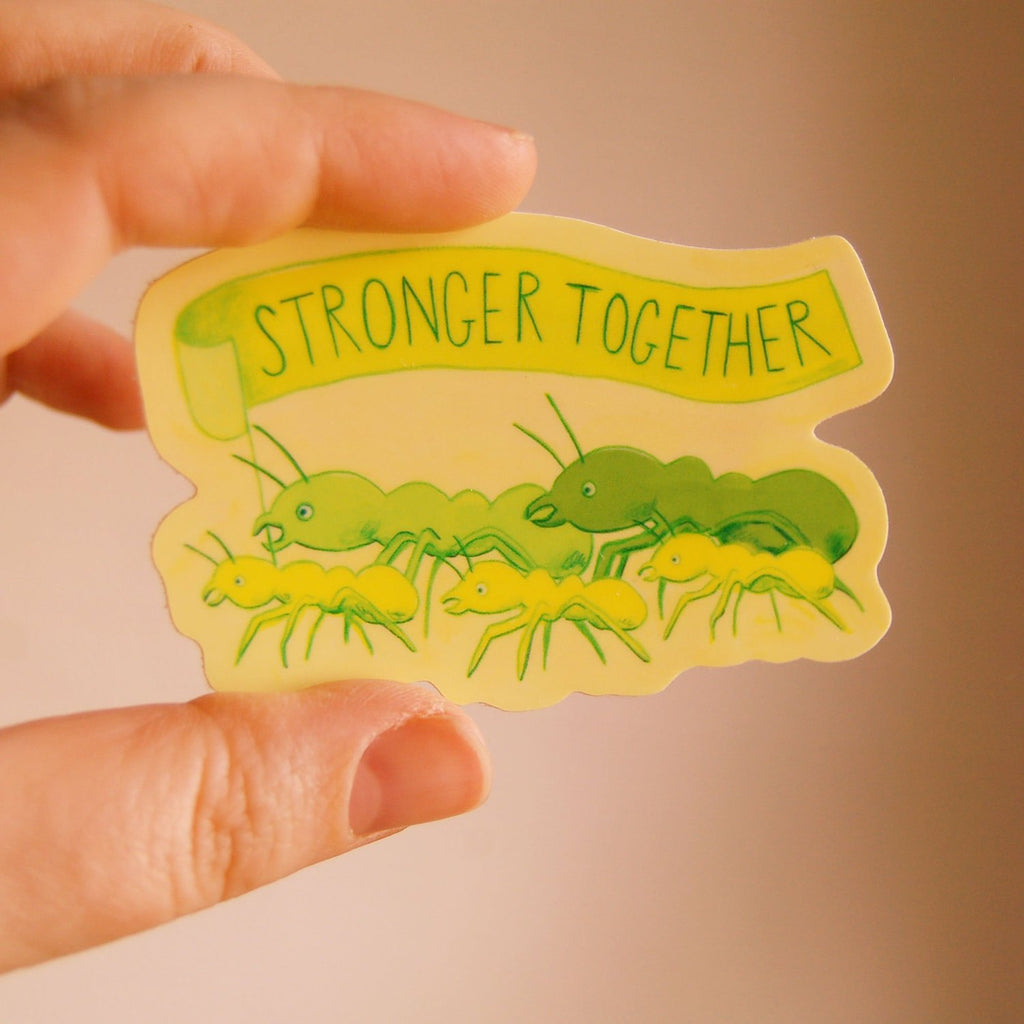 A hand holds a die-cut sticker featuring green ants carrying yellow banner that reads 'STRONGER TOGETHER' on a warm beige background.