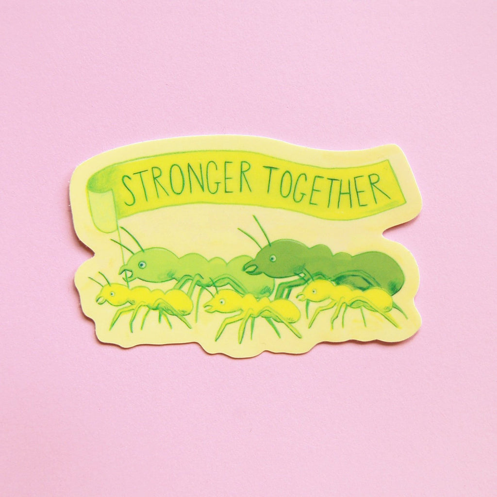 A die-cut sticker featuring a yellow banner that reads 'STRONGER TOGETHER' above green ants, shown on a solid pink background.