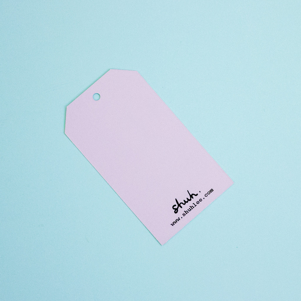 A lilac paper gift tag with a hanging hole and black printed text reading 'shuh.' and 'www.shuhlee.com'
