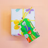 A square gift box wrapped in white paper with colourful abstract shapes, tied with a yellow ribbon and a matching illustrated paper tag, set against a soft peach background.