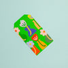 A green paper gift tag with a abstract floral pattern, shown diagonally on a solid light blue background.