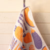 A white waffle-weave kitchen towel with a blue and orange vegetable pattern, hanging from a metal hook against a cream-coloured paneled wall.
