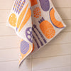 A textured kitchen towel with a vegetable motifs on a white background, hanging against a light beige tiled wall.