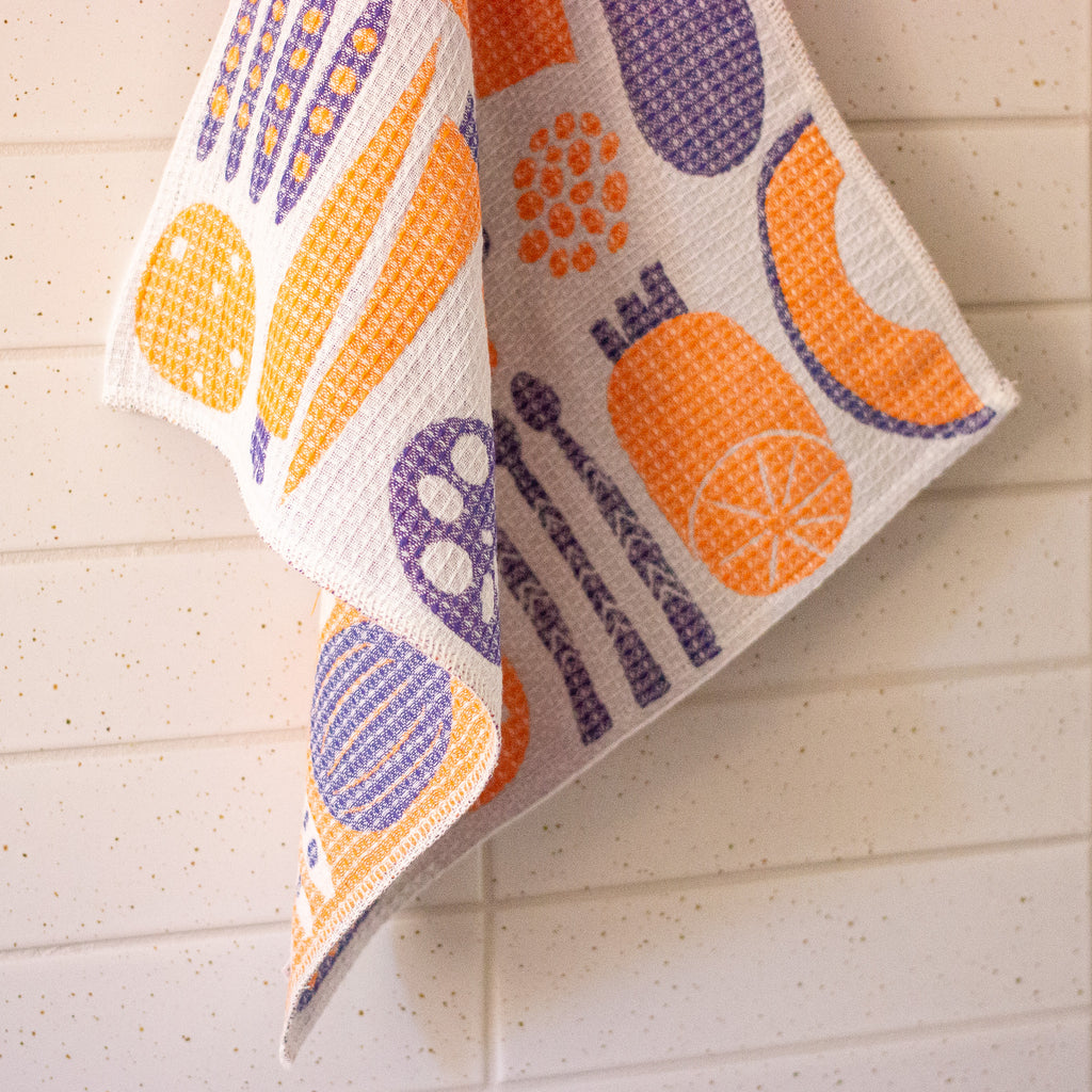 A textured kitchen towel with a vegetable motifs on a white background, hanging against a light beige tiled wall.