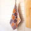 A textured kitchen towel with a colourful vegetable motifs like broccoli, pumpkin and peas pattern hangs from a metal hook on a white tiled wall next to a wooden cutting board.
