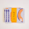 A folded Japanese dish towel with a white waffle-weave texture, featuring purple utensil patterns and an pumpkin design, wrapped with a bright orange paper band.