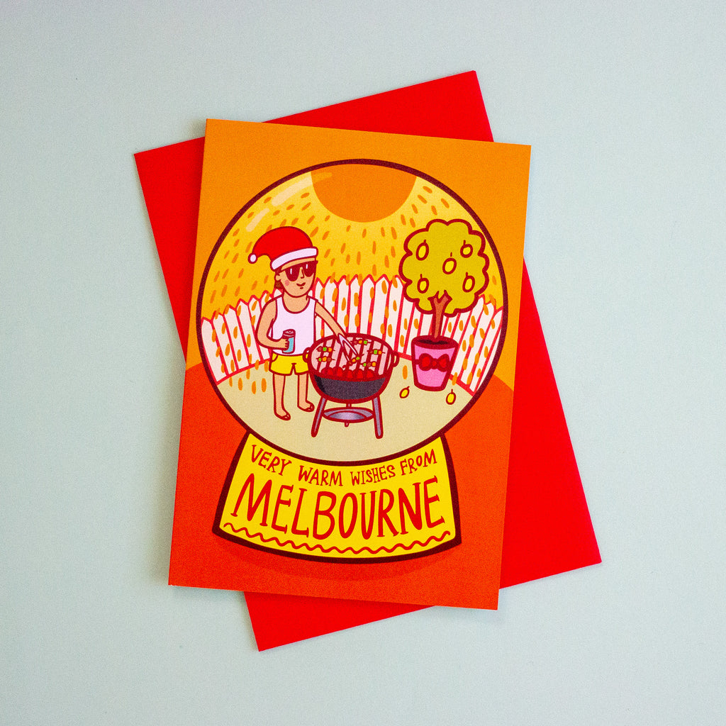 An A6 greeting card with a bright orange and red design, featuring an illustrated scene of a person grilling outdoors with the text 'VERY WARM WISSES FROM MELBOURNE', placed on top of a red envelope against a light grey background.