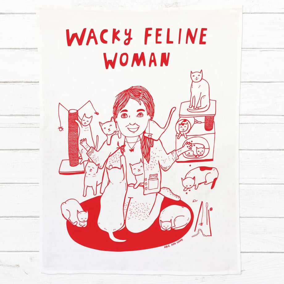 A white fabric tea towel featuring a red line art illustration of a woman surrounded by cats, with the text 'WACKY FELINE WOMAN' printed at the top, displayed flat against a white wood plank background.