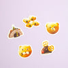 Five cute animal-shaped stickers with white borders arranged on a light lavender background. The designs include a brown bear wearing a green hat, a group of yellow koalas, a surprised yellow bear face, a yellow cat face, and a reindeer.