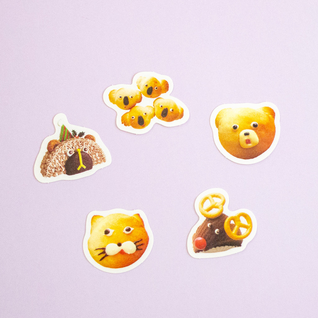 Five cute animal-shaped stickers with white borders arranged on a light lavender background. The designs include a brown bear wearing a green hat, a group of yellow koalas, a surprised yellow bear face, a yellow cat face, and a reindeer.