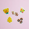 Five cute animal-shaped stickers arranged on a light pink background. The stickers feature a yellow bear wearing a green bow, two brown cats holding a heart, a yellow Christmas tree, a bunny and bear balloons, and three brown hedgehogs with black faces.