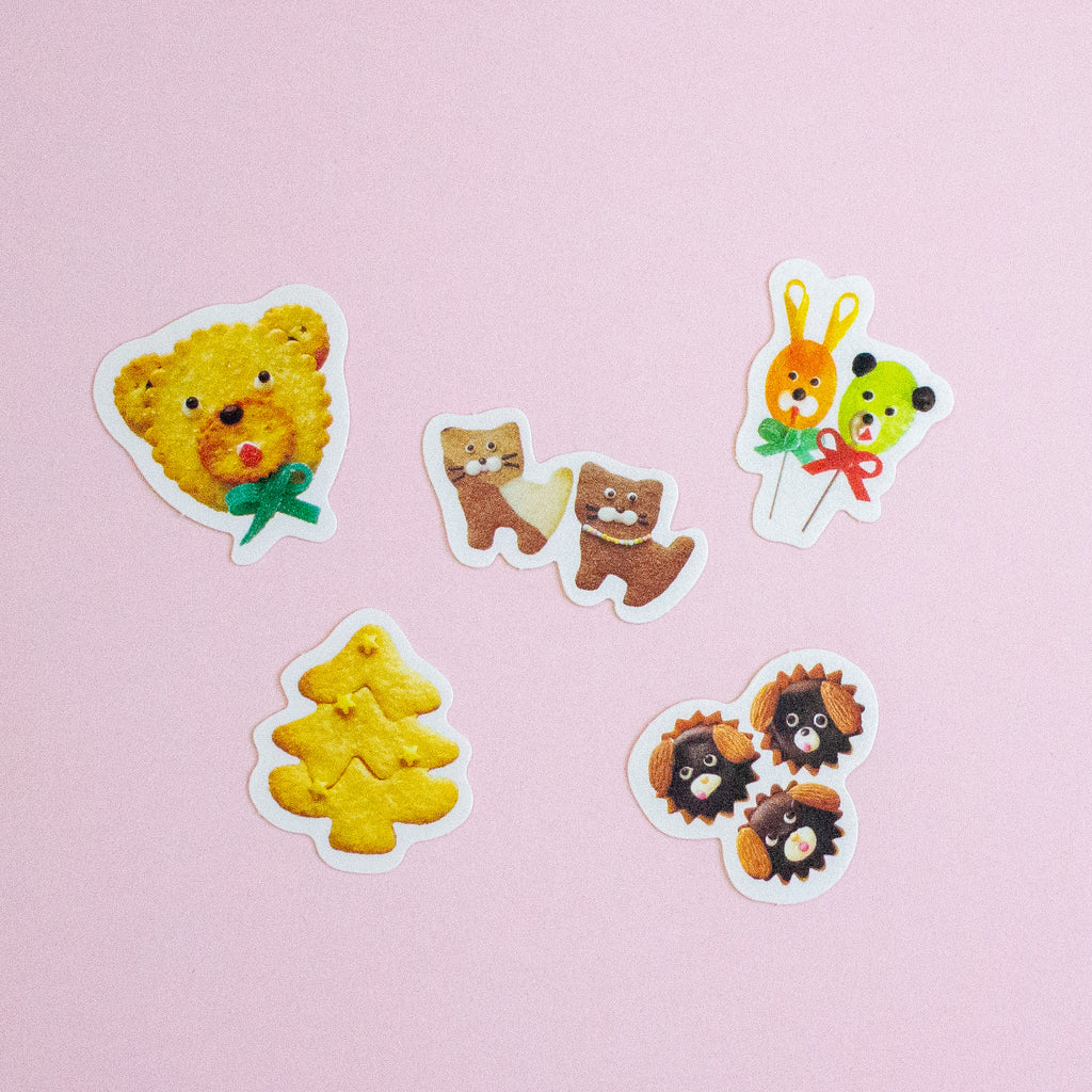 Five cute animal-shaped stickers arranged on a light pink background. The stickers feature a yellow bear wearing a green bow, two brown cats holding a heart, a yellow Christmas tree, a bunny and bear balloons, and three brown hedgehogs with black faces.