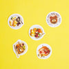 Five white plates with whimsical shapes and patterns, each decorated with colourful breakfast foods like pancakes, fruit, and eggs, arranged on a solid bright yellow background.