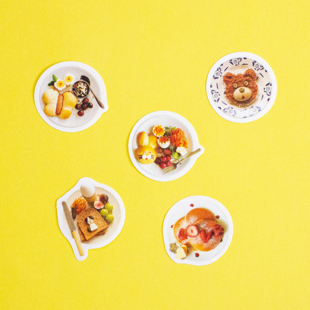 Five white plates with whimsical shapes and patterns, each decorated with colourful breakfast foods like pancakes, fruit, and eggs, arranged on a solid bright yellow background.