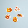 Five colourful food-themed stickers arranged on a light blue background, including a cat face, strawberries, a bear on a plate, and a bunny face.