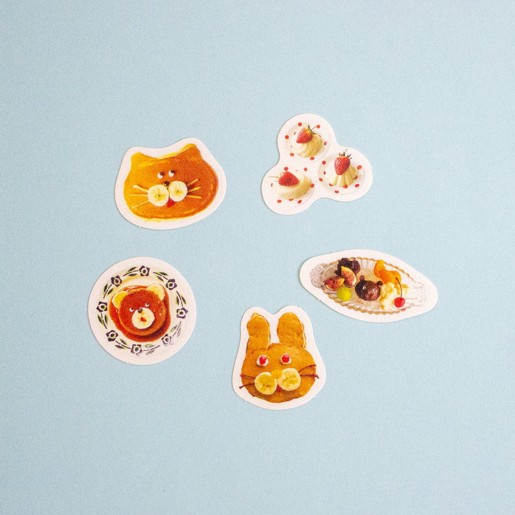 Five colourful food-themed stickers arranged on a light blue background, including a cat face, strawberries, a bear on a plate, and a bunny face.
