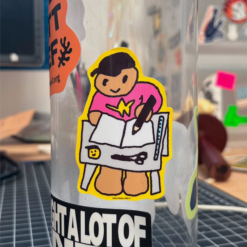A die-cut vinyl sticker of a cartoon artist drawing in a notebook, applied to a clear tumbler. The sticker features a character with black cap, a pink shirt with a yellow star, and tan legs, surrounded by a bright yellow border.